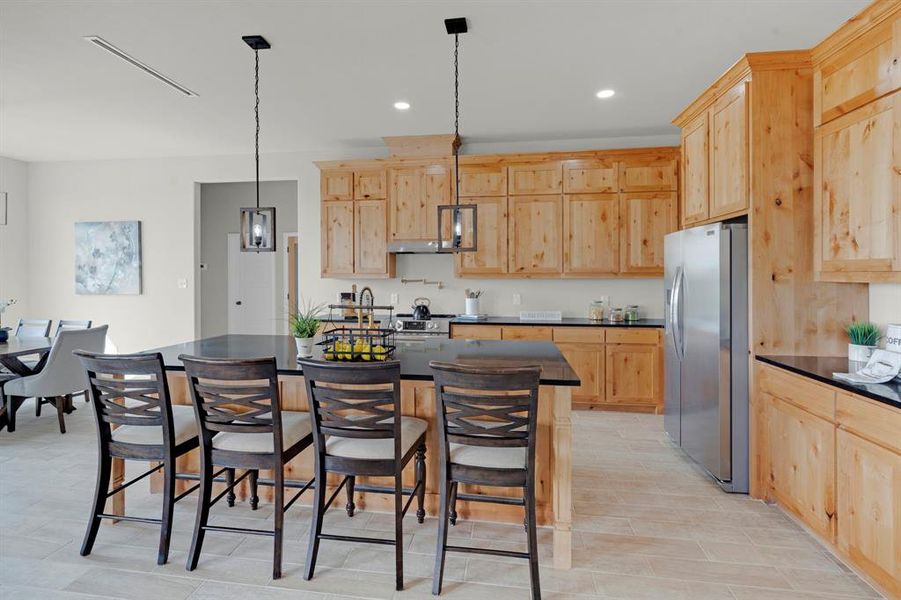 Kitchen with light wood-type flooring, stainless steel fridge with ice dispenser, light brown cabinets, pendant lighting, and a center island with sink