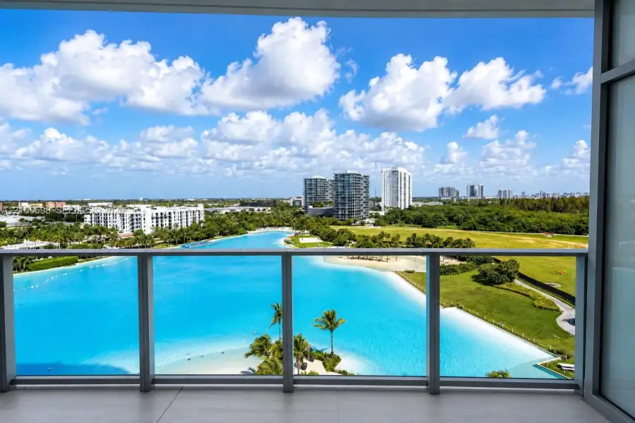 Covered balcony featuring a glass and metal railing, overlooking an expansive lagoon, sandy shoreline, and palm trees