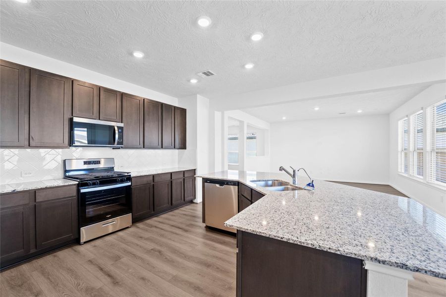 This spacious kitchen boasts soaring ceilings, Woodmont Dakota cabinets in a Smoke stain, Granite Luna Pearl countertops, stainless steel Whirlpool appliances, a modern Daltile backsplash, recessed lighting, an extended breakfast bar countertop, and a pantry—all overlooking the expansive family room and breakfast nook.