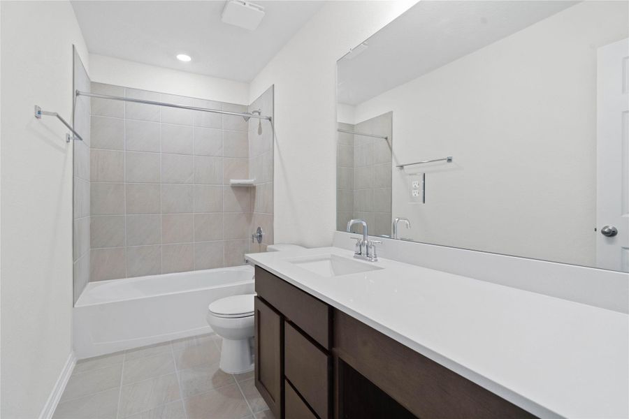 The secondary bathroom offers tile flooring, a bath/shower combination with a stylish tile surround, rich stained wood cabinetry, elegant light countertops, a mirror, and sleek fixtures with modern finishes.