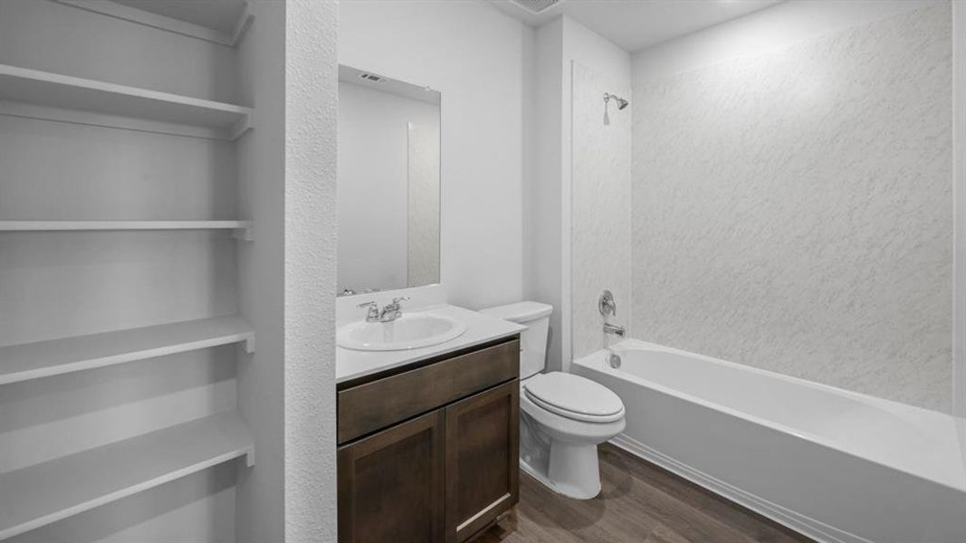 Bathroom featuring built-in shelving, wood-finish flooring, a white vanity with an integrated sink, a toilet, and a bathtub with a textured shower surround