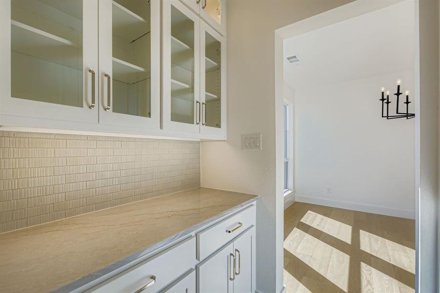 Bar featuring tasteful backsplash, white cabinets, light stone countertops, glass insert cabinets, and light wood-type flooring Bar featuring tasteful backsplash, white cabinets, light stone countertops, glass insert cabinets, and light wood-type flooring