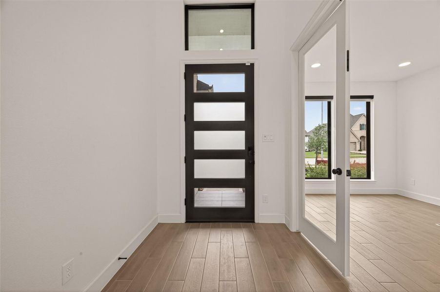 This entryway features a modern, frosted glass door, wood-look tile flooring, and leads to a bright room with large windows, offering a welcoming and stylish first impression.