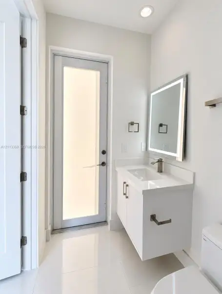Floating vanity with white cabinetry, rectangular basin, integrated countertop, and brushed metal hardware, complemented by a backlit rectangular mirror, polished tile flooring, and a glass-panel exterior door