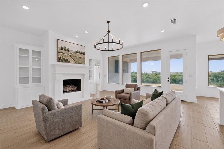 Spacious living area featuring a central fireplace with mantle, built-in cabinetry, wood-finish flooring, a contemporary chandelier, and large windows with outdoor views Spacious living area featuring a central fireplace with mantle, built-in cabinetry, wood-finish flooring, a contemporary chandelier, and large windows with outdoor views