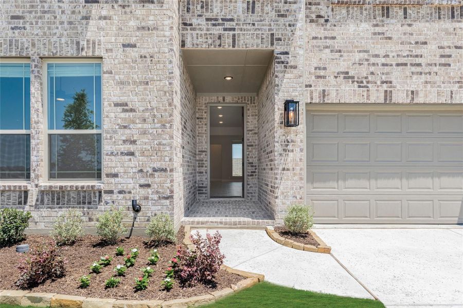 Home entrance with light brick exterior, covered front door, wall lantern, and two-car garage beside landscaped flower bed.