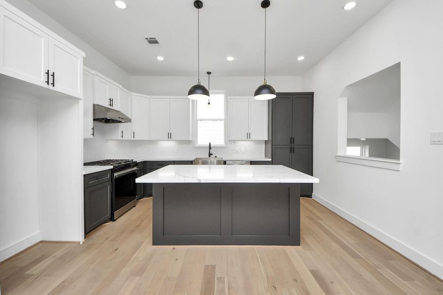 This modern kitchen features a spacious island with a white countertop, sleek black lower cabinets, and contrasting white upper cabinets. It boasts stainless steel appliances, pendant lighting, and light wood flooring, creating a bright and inviting space.
