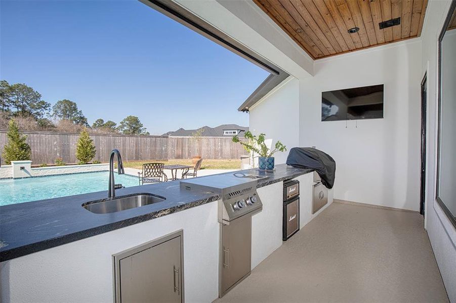 This photo showcases a modern outdoor kitchen with a sink, grill, and mini fridge, overlooking a backyard with a pool. The area features a wooden ceiling and a mounted TV, perfect for entertaining.