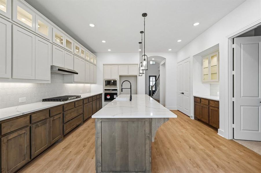 Kitchen featuring glass insert cabinets, an island with sink, arched walkways, backsplash, and hanging light fixtures Kitchen featuring glass insert cabinets, an island with sink, arched walkways, backsplash, and hanging light fixtures