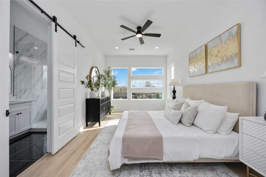 This impressive primary bedroom offers a bright, airy layout enhanced by expansive modern windows that frame beautiful outdoor views, warm wood-look flooring, recessed lighting, and a sleek ceiling fan; a contemporary sliding barn door opens to the luxurious en-suite bathroom, while elegant staging and soft neutral tones highlight the room’s spacious and inviting design.