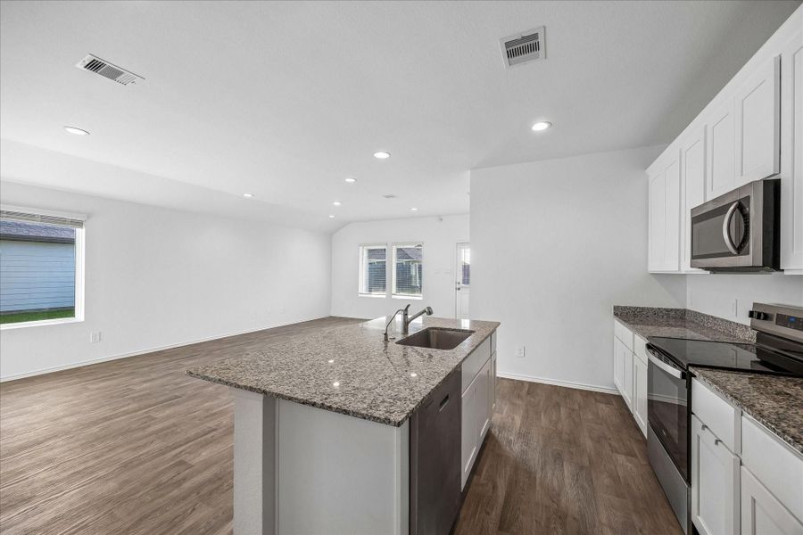 Spacious, open-concept kitchen and living area. The kitchen features granite countertops, a large island with a sink, stainless steel appliances, and white cabinetry. The space is well-lit with recessed lighting and has wood-look flooring, creating a modern and inviting atmosphere.