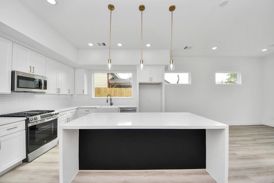 This modern kitchen features sleek white cabinetry, stainless steel appliances, and a large island with pendant lighting. The open layout and light wood flooring create a bright and inviting space.