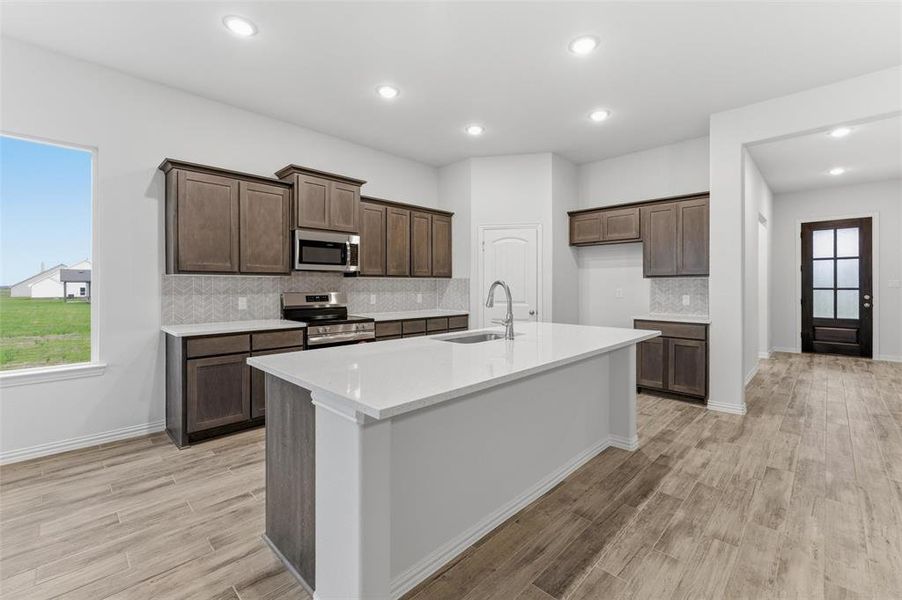Modern kitchen featuring wood-finish flooring, recessed lighting, an island with an integrated sink, dark wood cabinetry, and stainless steel appliances
