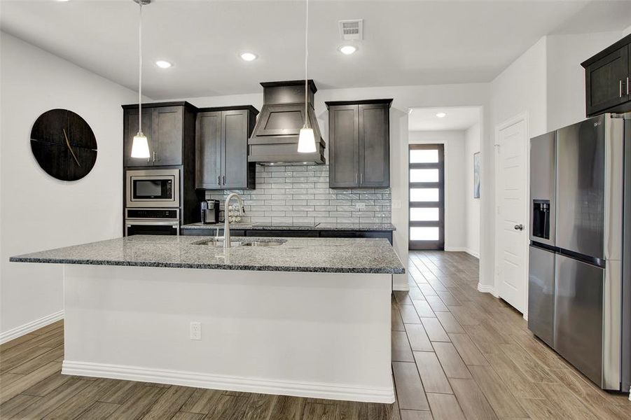 Kitchen with light hardwood / wood-style flooring, an island with sink, stainless steel appliances, custom exhaust hood, and decorative backsplash