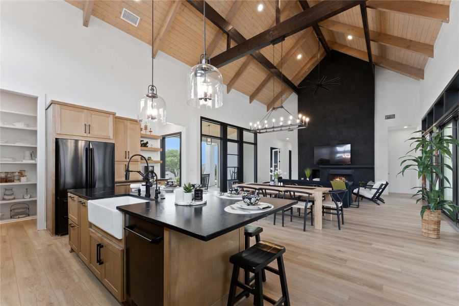 Open-concept living space featuring vaulted wood-plank ceilings with exposed beams, light wood-finish flooring, and a kitchen island with a farmhouse sink and dark countertop Open-concept living space featuring vaulted wood-plank ceilings with exposed beams, light wood-finish flooring, and a kitchen island with a farmhouse sink and dark countertop