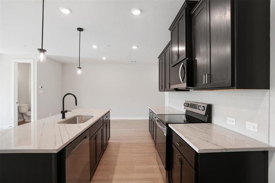 Kitchen with dark cabinets, stainless steel appliances, light stone counters, pendant lighting, and light wood-style flooring Kitchen with dark cabinets, stainless steel appliances, light stone counters, pendant lighting, and light wood-style flooring