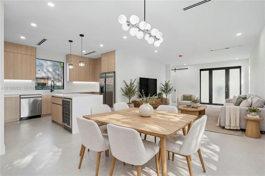 Open-concept kitchen featuring light wood-finish cabinetry, a central island with an integrated beverage cooler, stainless steel appliances, and a modern multi-globe chandelier Open-concept kitchen featuring light wood-finish cabinetry, a central island with an integrated beverage cooler, stainless steel appliances, and a modern multi-globe chandelier