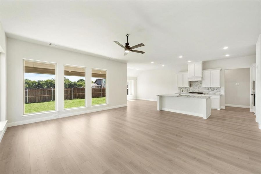Open-concept living space featuring wood-finish flooring, a contemporary ceiling fan, and three large windows overlooking a fenced backyard Open-concept living space featuring wood-finish flooring, a contemporary ceiling fan, and three large windows overlooking a fenced backyard