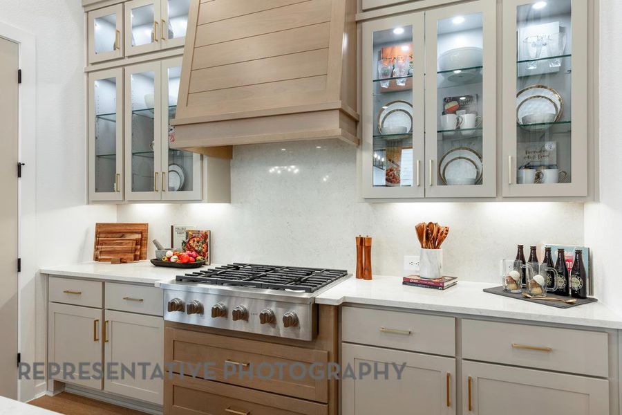 Kitchen featuring light stone counters, stainless steel gas stovetop, custom range hood, tasteful backsplash, and glass insert cabinets