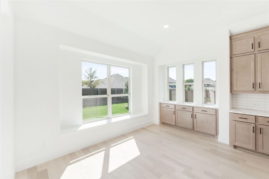 Bright dining area with light wood floors, tan cabinets, and large windows overlooking a fenced backyard