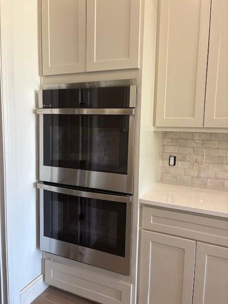 Kitchen view of stainless steel double oven, light stone counters, white cabinetry, and tasteful backsplash