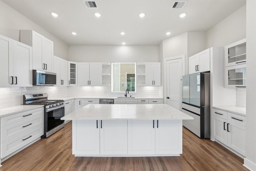 Bright, modern chef’s kitchen featuring a large quartz island, stainless steel appliances, shaker cabinetry, and subway tile backsplash under recessed lighting.