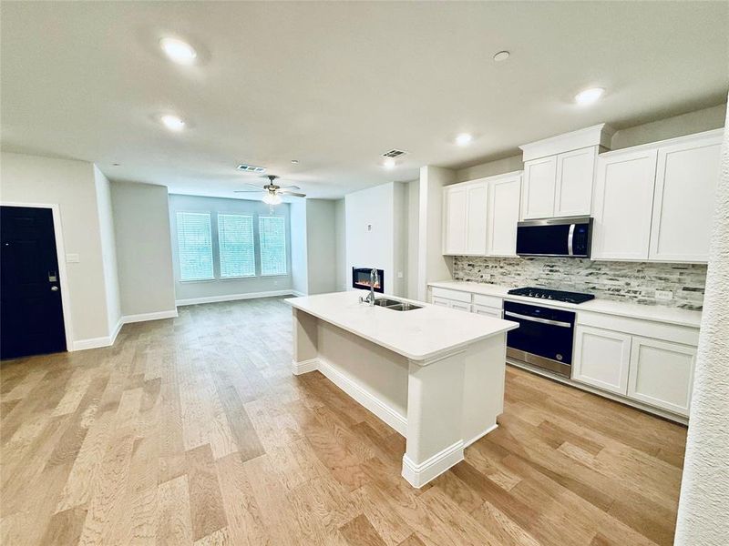 Open-concept living space featuring wood-finish flooring, a kitchen island with integrated sink, white cabinetry, a tiled backsplash, and recessed lighting