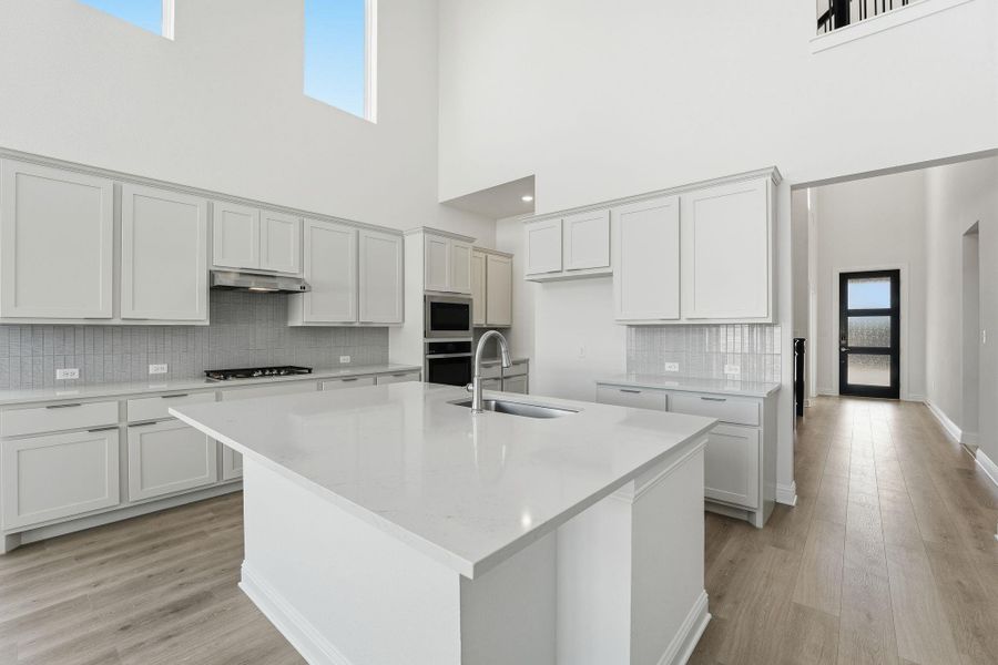 Kitchen featuring decorative backsplash, light stone counters, a kitchen island with sink, light wood-type flooring, and a towering ceiling