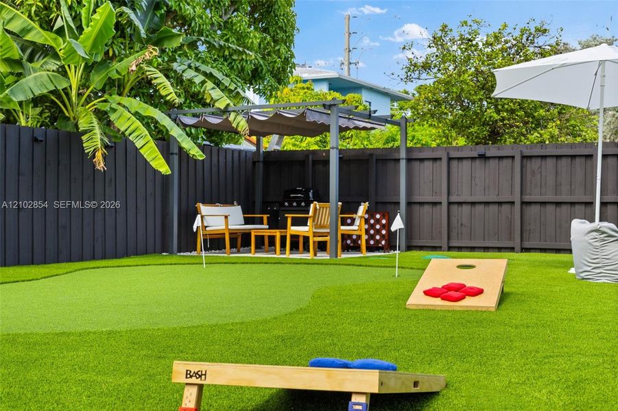 Fenced backyard featuring synthetic turf, a built-in putting green, and a metal-frame pergola with retractable canopy