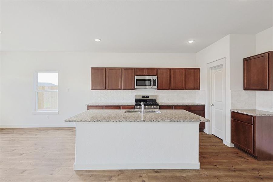 Kitchen with stainless steel appliances, light stone counters, backsplash, a kitchen island with sink, and light wood-type flooring