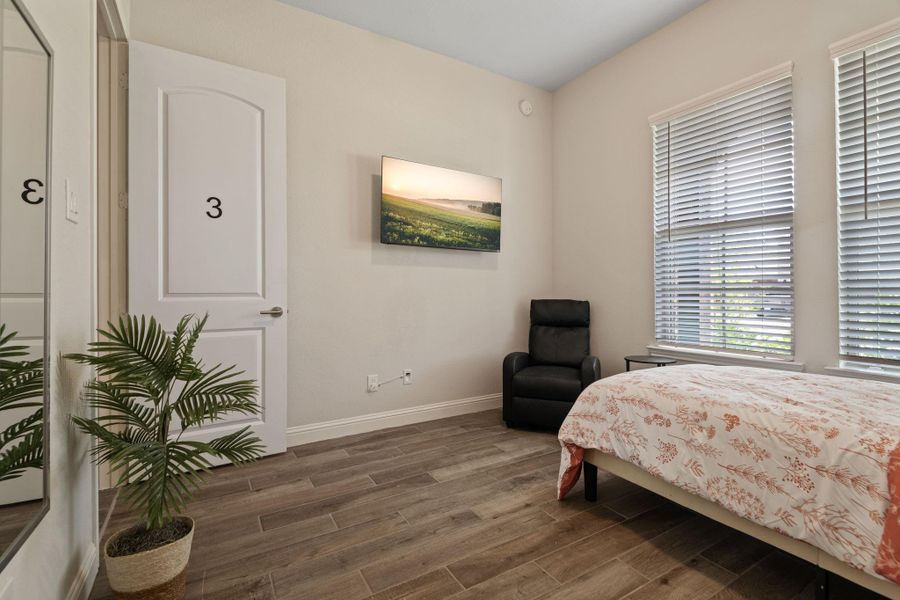 This cozy bedroom features wood-style flooring, a comfortable bed with floral bedding, and a small recliner. It has ample natural light from two large windows with blinds, a mirrored closet door, and a simple artwork on the wall. This cozy bedroom features wood-style flooring, a comfortable bed with floral bedding, and a small recliner. It has ample natural light from two large windows with blinds, a mirrored closet door, and a simple artwork on the wall.