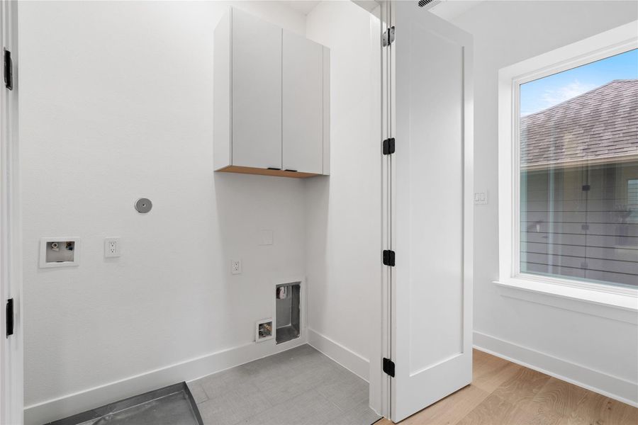 Utility, elevated: a dedicated laundry room with floor tile underfoot, fitted upper cabinetry for concealed storage, and smart hookups for both gas and electric. A high window washes the space in daylight while maintaining privacy, turning everyday tasks into a clean, well-composed ritual