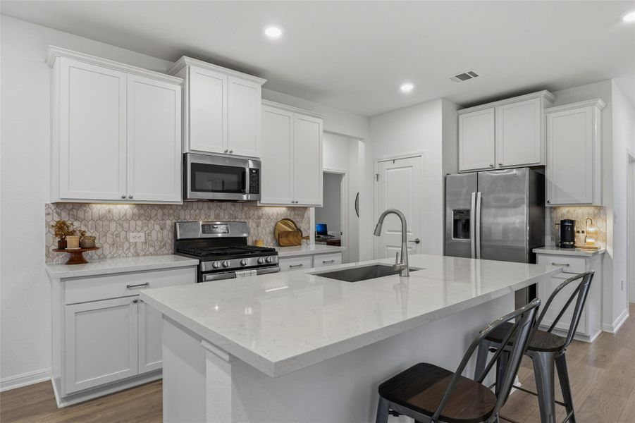 Kitchen featuring white shaker-style cabinetry, a large center island with a sink, stainless steel appliances, a geometric tile backsplash, and light wood-finish flooring
