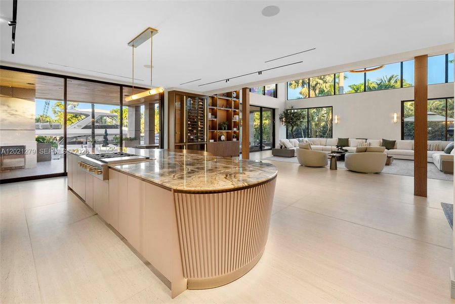 Wide-angle view of open-plan kitchen and great room with curved fluted island, book-matched stone countertop, brass cooktop fixtures, full-height illuminated shelving, floor-to-ceiling glass with canal and yacht views, and wraparound sectional seating beyond.