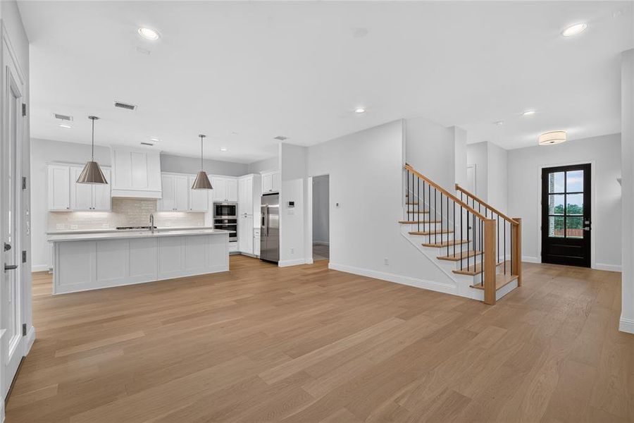 Open-concept living space featuring wood-finish flooring, a contemporary kitchen with white cabinetry and stainless steel appliances, a central island with pendant lighting, a staircase with wood treads and iron balusters, and a dark-paneled door with glass inserts Open-concept living space featuring wood-finish flooring, a contemporary kitchen with white cabinetry and stainless steel appliances, a central island with pendant lighting, a staircase with wood treads and iron balusters, and a dark-paneled door with glass inserts