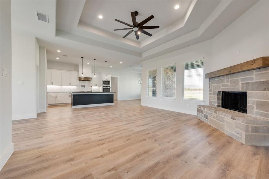 Open-concept living space featuring wood-finish flooring, a stone fireplace with a wood mantle, and a tray ceiling with recessed lighting and a ceiling fan