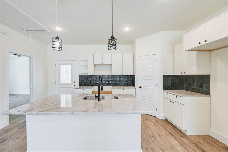 Kitchen featuring a spacious island with an undermount sink and a matte black faucet, white cabinetry, dark subway tile backsplash, wood-finish flooring, and two pendant lights Kitchen featuring a spacious island with an undermount sink and a matte black faucet, white cabinetry, dark subway tile backsplash, wood-finish flooring, and two pendant lights
