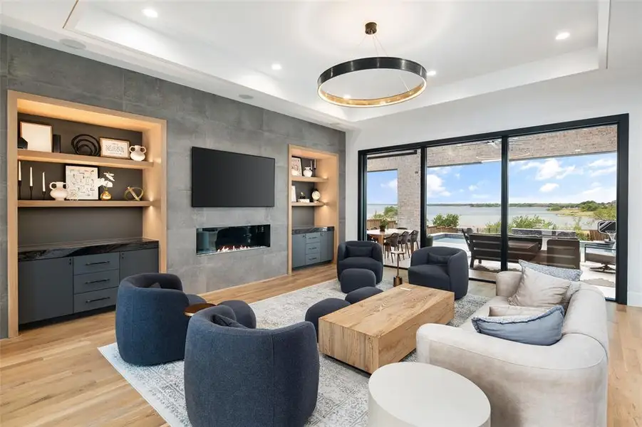 Elegant living room designed for both comfort and style, featuring a tray ceiling with accent lighting that adds depth and architectural interest. Custom built-in shelving with accent lighting and cabinetry provide both warmth and function, while expansive sliding glass doors frame stunning lake views and fill the space with natural light.