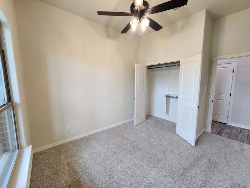 Carpeted room featuring a ceiling fan with light fixture, double-door closet with shelving, large window with sill, and neutral wall tones