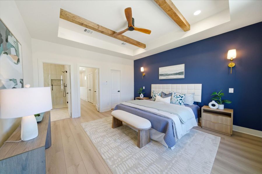This spacious bedroom features a modern design with a bold blue accent wall, wooden ceiling beams, and a ceiling fan. It includes an ensuite bathroom with a glass shower, stylish lighting, and a cozy seating area. The space is bright and inviting, ideal for relaxation.