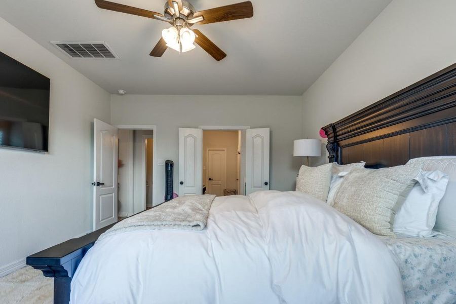 This bedroom features a cozy setup with a large bed,  A ceiling fan provides comfort, while a mounted TV offers entertainment. The room is well-lit with a lamp, and multiple doors provide easy access to adjoining spaces.