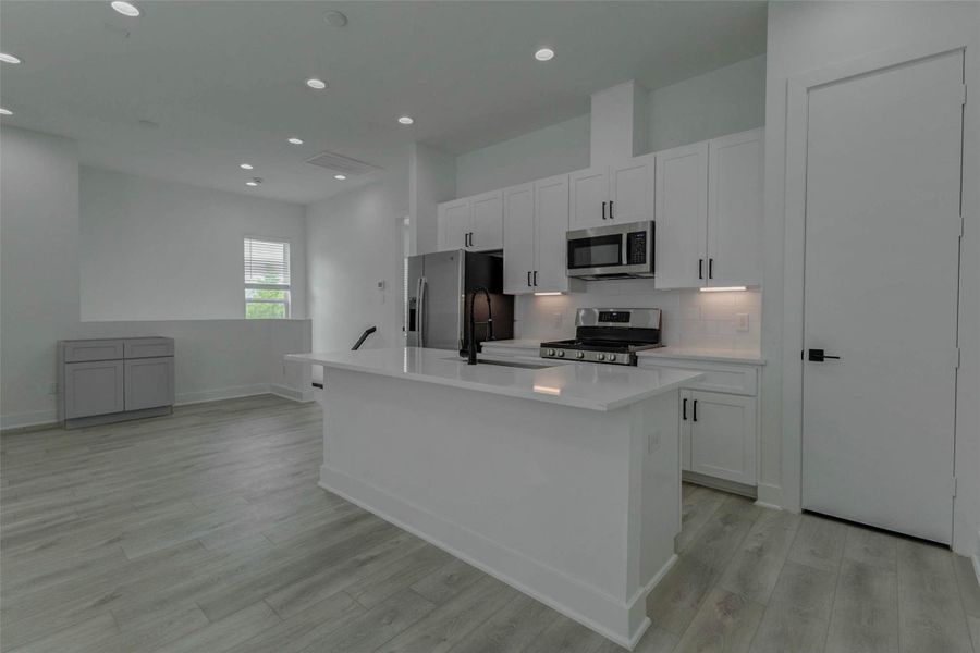 Modern kitchen view featuring a large white island, quartz-style countertops, stainless appliances, gas range, built-in microwave, white shaker-style cabinetry, black hardware, and under-cabinet lighting. The open layout flows into the living area, creating a bright, contemporary space for cooking, dining, entertaining, and everyday living.