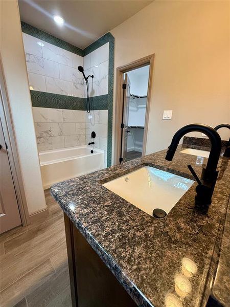 Bathroom featuring a dual vanity with granite countertops and undermount sinks, wood-finish flooring, and a tub/shower combination with tile surround and dark-finish fixtures Bathroom featuring a dual vanity with granite countertops and undermount sinks, wood-finish flooring, and a tub/shower combination with tile surround and dark-finish fixtures
