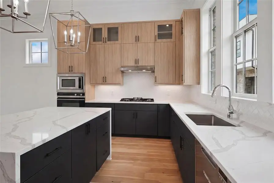 Contemporary kitchen featuring a large central island, light wood-finish upper cabinetry, matte black lower cabinetry, stainless steel appliances, and white countertops with subtle veining