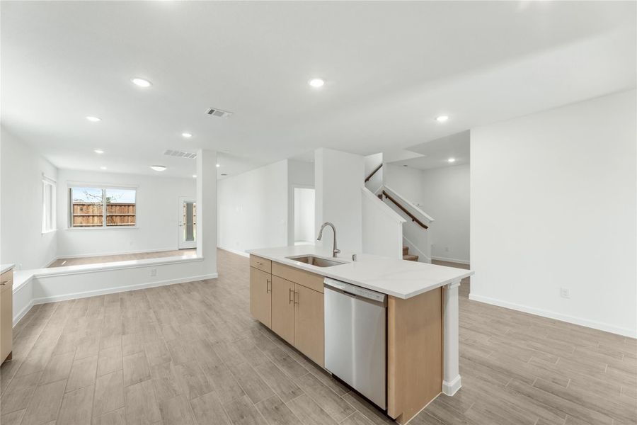 Kitchen island with sink, dishwasher, and light wood cabinets in open-concept living area with wood-look floors