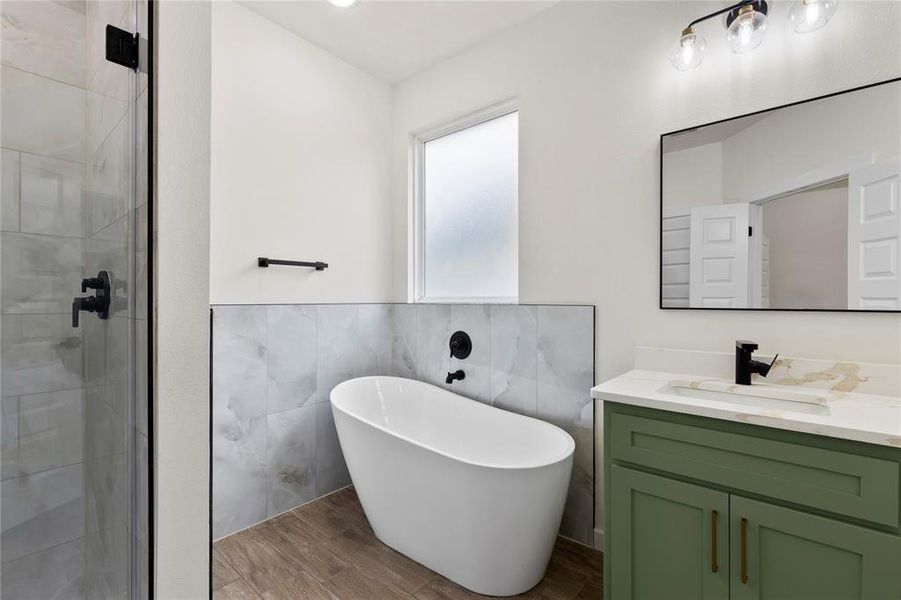Bathroom featuring a freestanding tub, glass-enclosed shower, wood-finish flooring, a single vanity with a green base, and a rectangular framed mirror