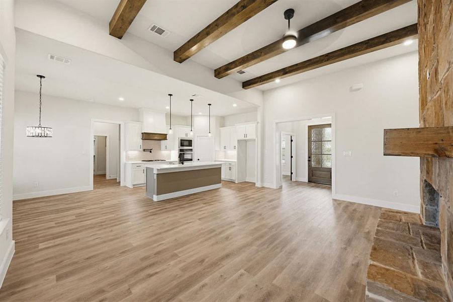 Vaulted ceilings with exposed wood beams, wood-finish flooring, a stone fireplace with a wood mantle, a kitchen island with pendant lighting, and white cabinetry