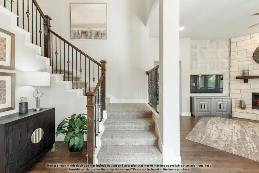 Carpeted staircase with wood and iron railing next to a living room with stone fireplace and hardwood floors