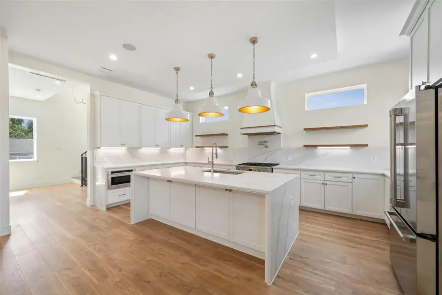 Kitchen features open layout with a large island, white cabinetry, pot filler and stainless steel appliances. It has wood flooring, pendant lighting, and ample natural light from high windows.