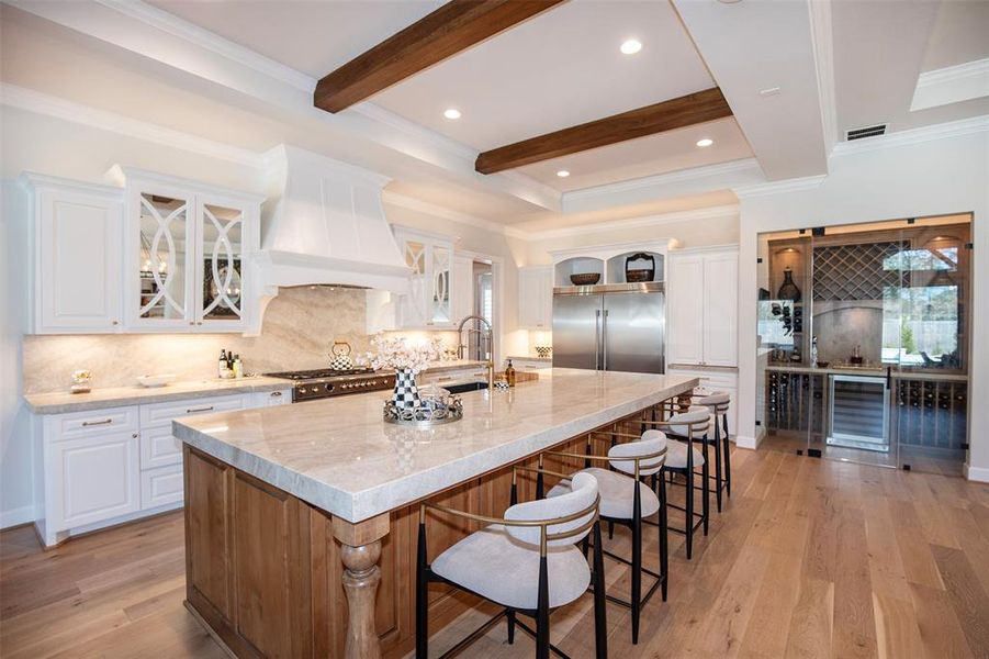 This kitchen boasts a spacious island with seating, elegant marble countertops, and high-end stainless steel appliances. It features white cabinetry, wooden ceiling beams, and a built-in wine storage area, creating a blend of modern and rustic charm.